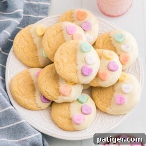 Vanilla cookies dipping in white chocolate and topped with 3 conversation heart candies. The Valentines cookies are served stacked on a white plate with a glass of milk, linen, and bowl of conversation hearts around them.
