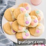 Vanilla cookies dipping in white chocolate and topped with 3 conversation heart candies. The Valentines cookies are served stacked on a white plate with a glass of milk, linen, and bowl of conversation hearts around them.