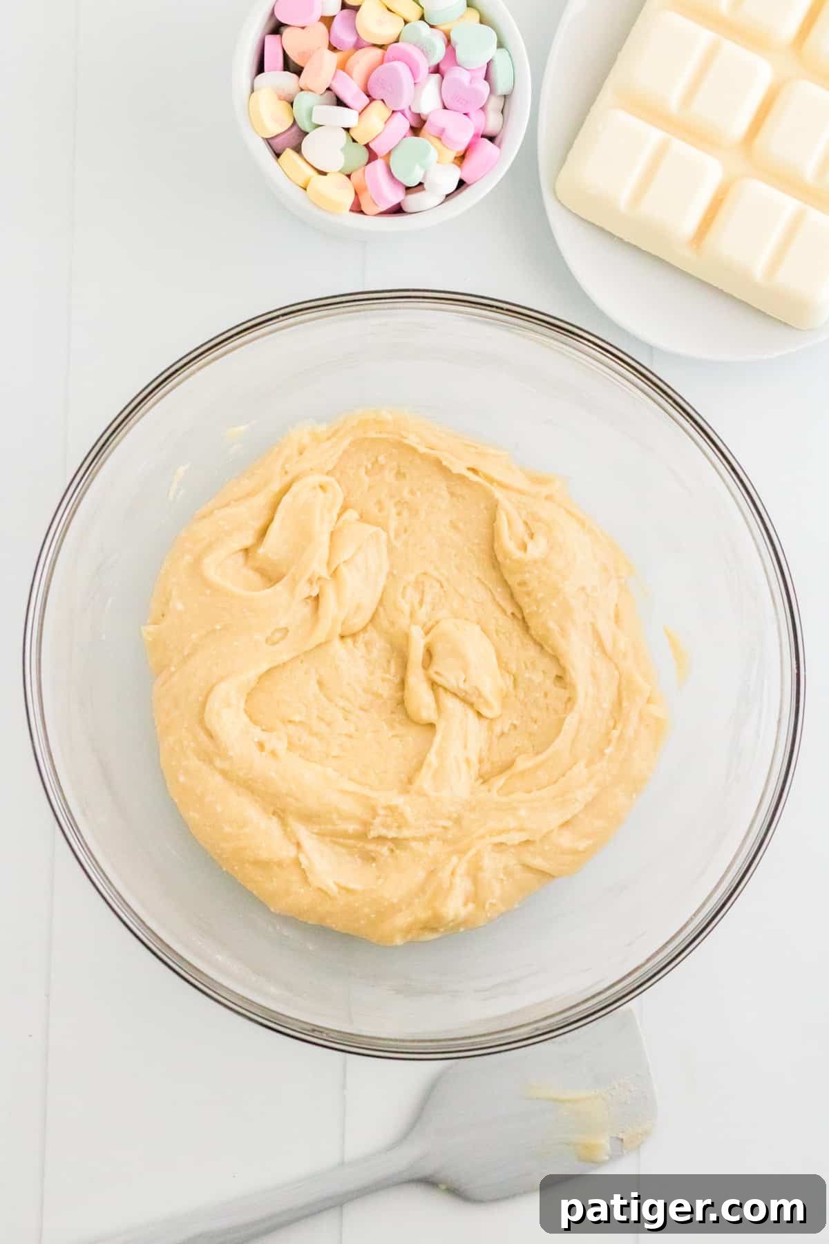 cookie batter in glass bowl with spatula, bowl or conversation heart candies, and almond bark around it