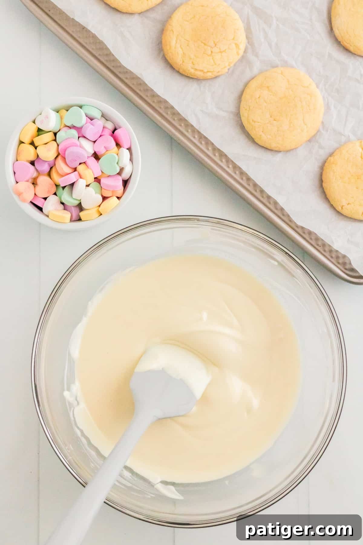 Melted almond bark in glass bowl with candy hearts and vanilla cookies on lined baking sheet in background
