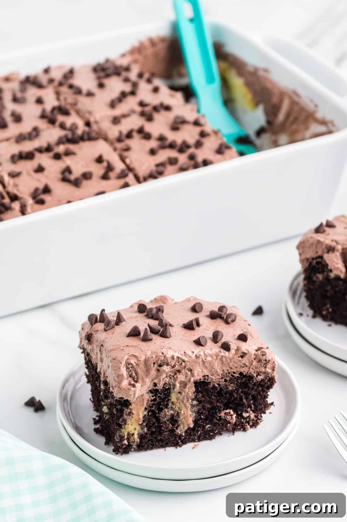 A delectable slice of Irish Cream Chocolate Poke Cake, featuring layers of moist chocolate cake, creamy filling, and a light chocolate frosting, garnished with chocolate shavings, served on a white plate with the rest of the cake in the background.