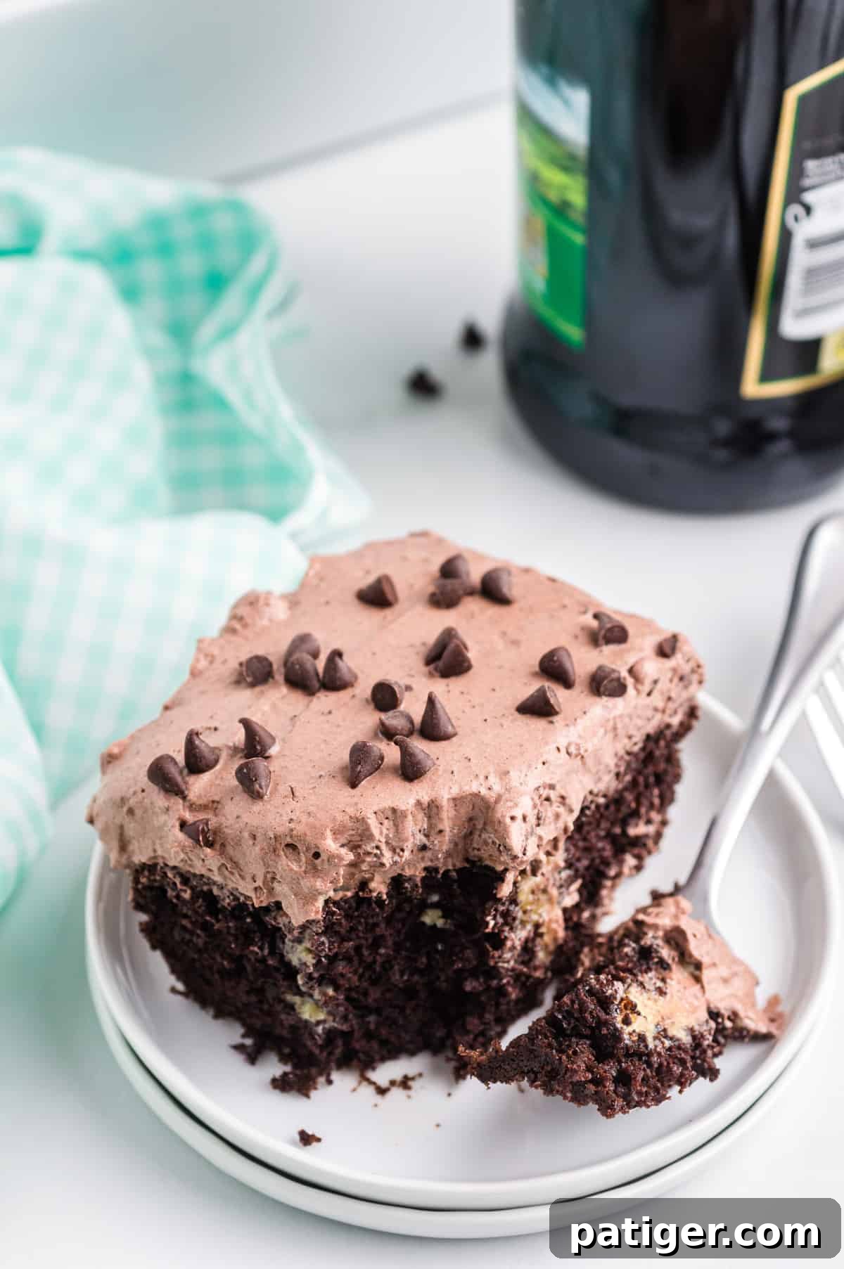 A close-up shot of a single slice of Baileys chocolate poke cake, elegantly presented on a white plate. The cake is topped with light and fluffy chocolate frosting and mini chocolate chips, with a small bite-size piece already taken out by a fork resting on the plate. A bottle of Baileys Irish Cream stands artfully blurred in the background, reinforcing the cake's key ingredient.
