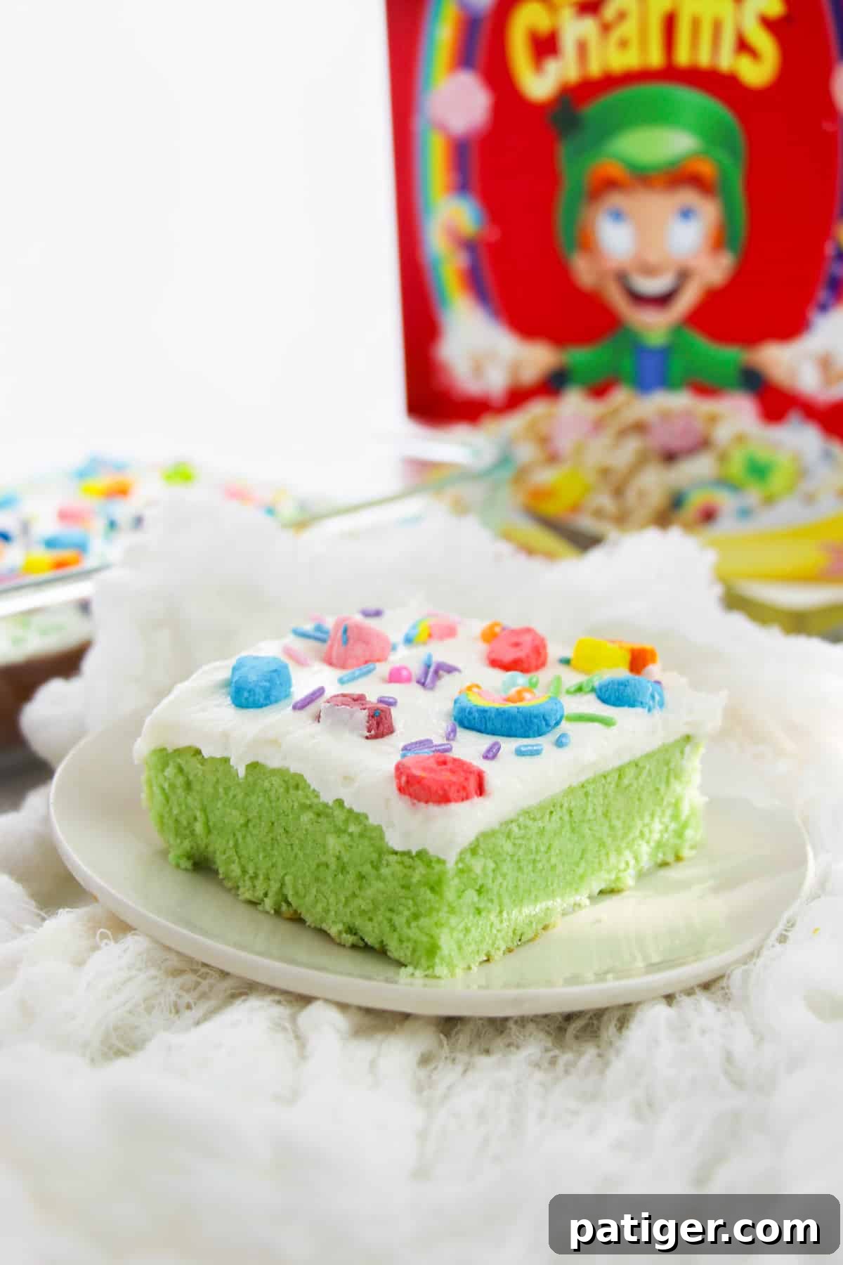 Green Lucky charms cake with white buttercream frosting, rainbow sprinkles, and lucky charms marshmallows on top. Box of Lucky Charms cereal is behind the slice of cake. 