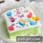Slice of lucky charms cereal cake on white plate. The cake is bright green with white frosting topped with rainbow sprinkles and lucky charms marshmallows.