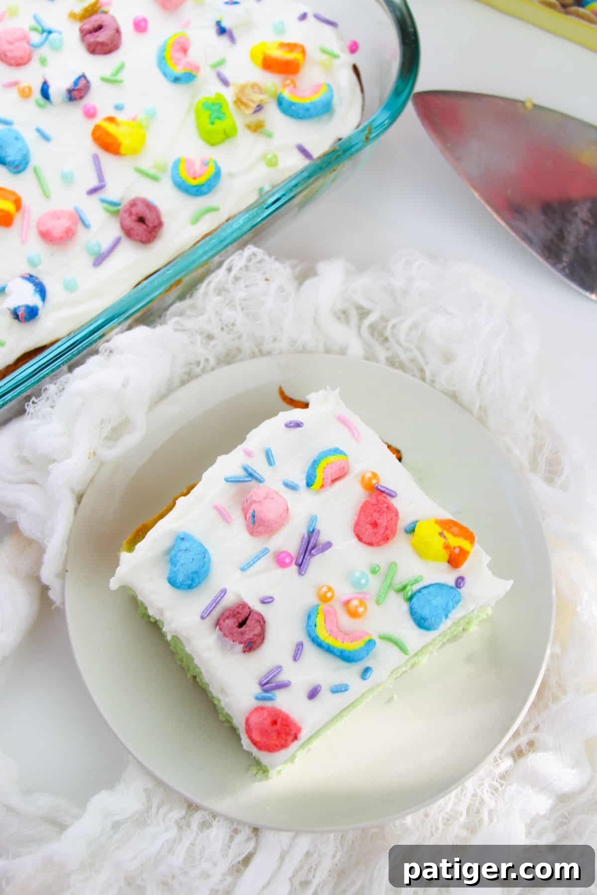 Slice of lucky charms sheet cake with white frosting topped with rainbow sprinkles and lucky charms marshmallows. Whole cake in baking pan can be seen next to the slice.