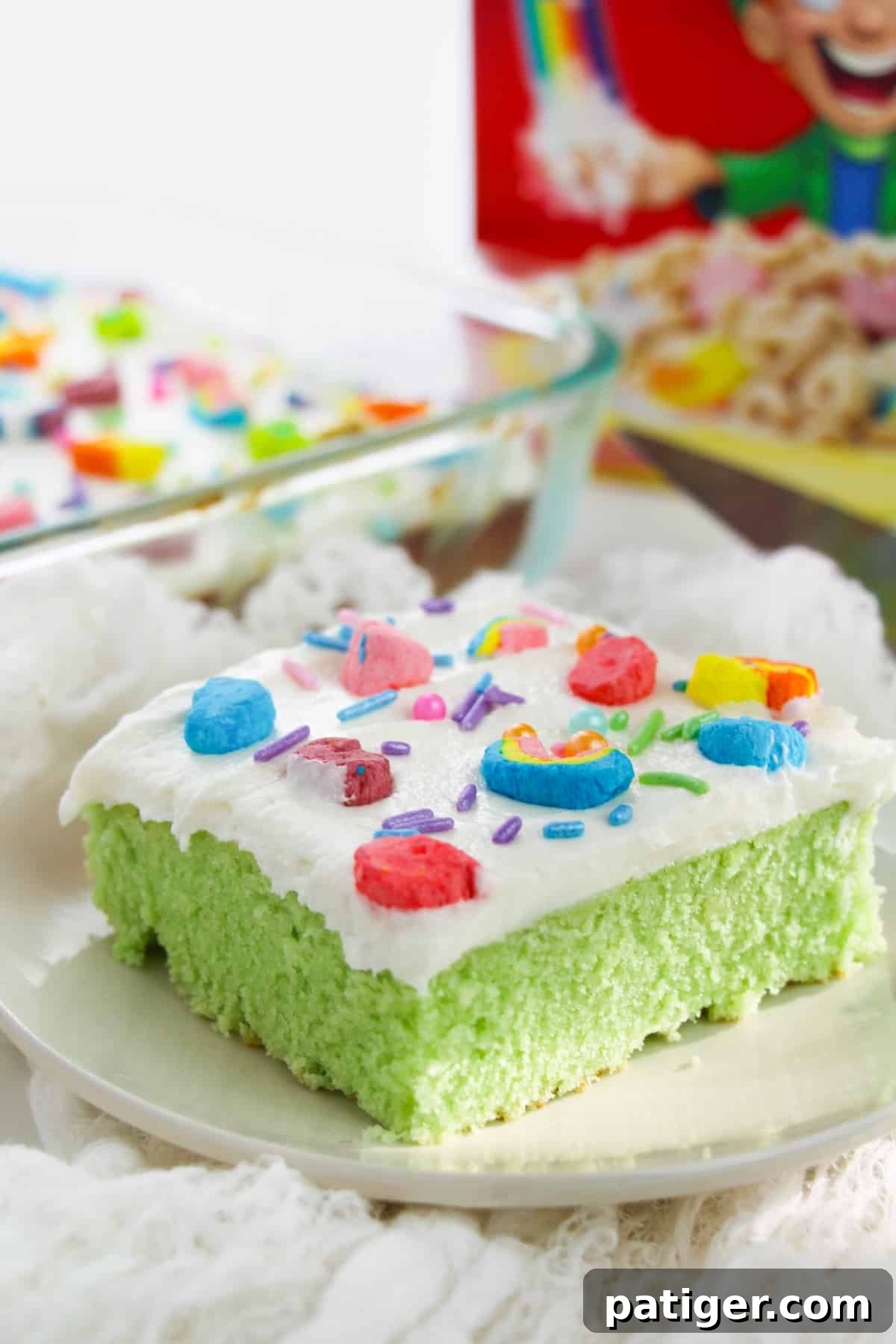 Green Lucky charms cake with white buttercream frosting, rainbow sprinkles, and lucky charms marshmallows on top. Box of Lucky Charms cereal is behind the slice of cake.