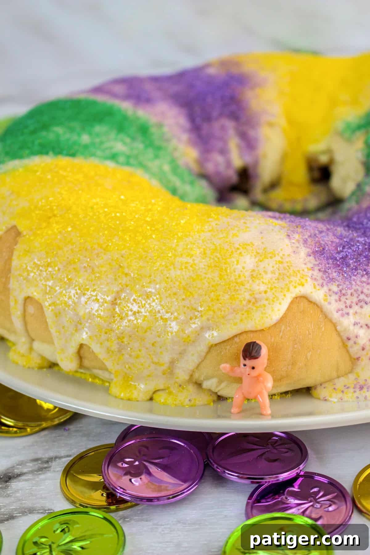 Easy homemade king cake with vanilla glaze and decorated with purple, green, and yellow sanding sugar. A small plastic baby in one the white platter next to the cake.