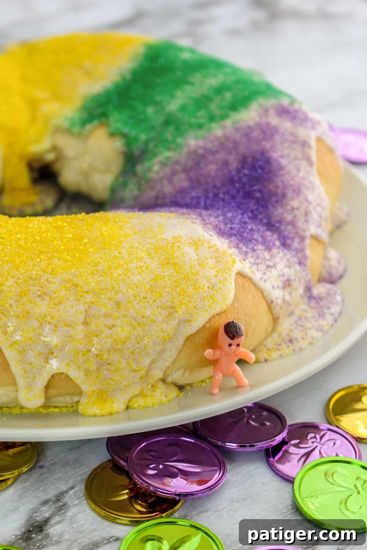 King cake with glaze and green, yellow, and purple sanding sugars. A small toy baby is leaning on the cake.