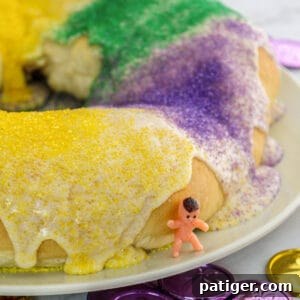 King cake with glaze and green, yellow, and purple sanding sugars. A small toy baby is leaning on the cake.