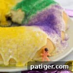 King cake with glaze and green, yellow, and purple sanding sugars. A small toy baby is leaning on the cake.
