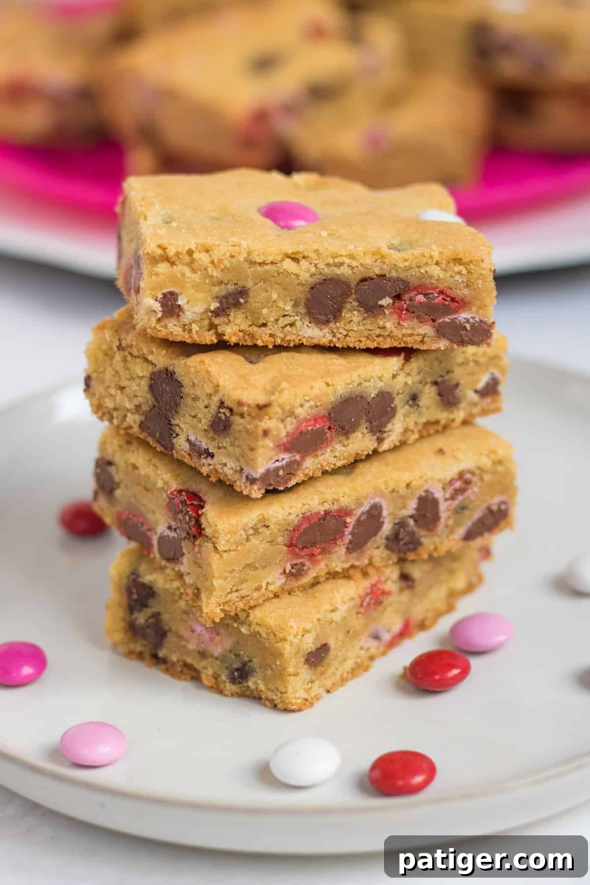 Four Valentine cookie bars with M&Ms stacked on top of one another on white plate with additional M&Ms sprinkled around them and more bars on bright pink plate in background.