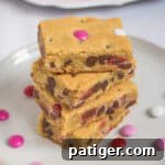 Four Valentine cookie bars with M&Ms stacked on top of one another on white plate with additional M&Ms sprinkled around them and more bars on bright pink plate in background.
