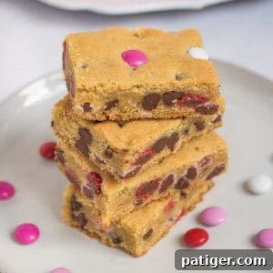 Four Valentine cookie bars with M&Ms stacked on top of one another on white plate with additional M&Ms sprinkled around them and more bars on bright pink plate in background.