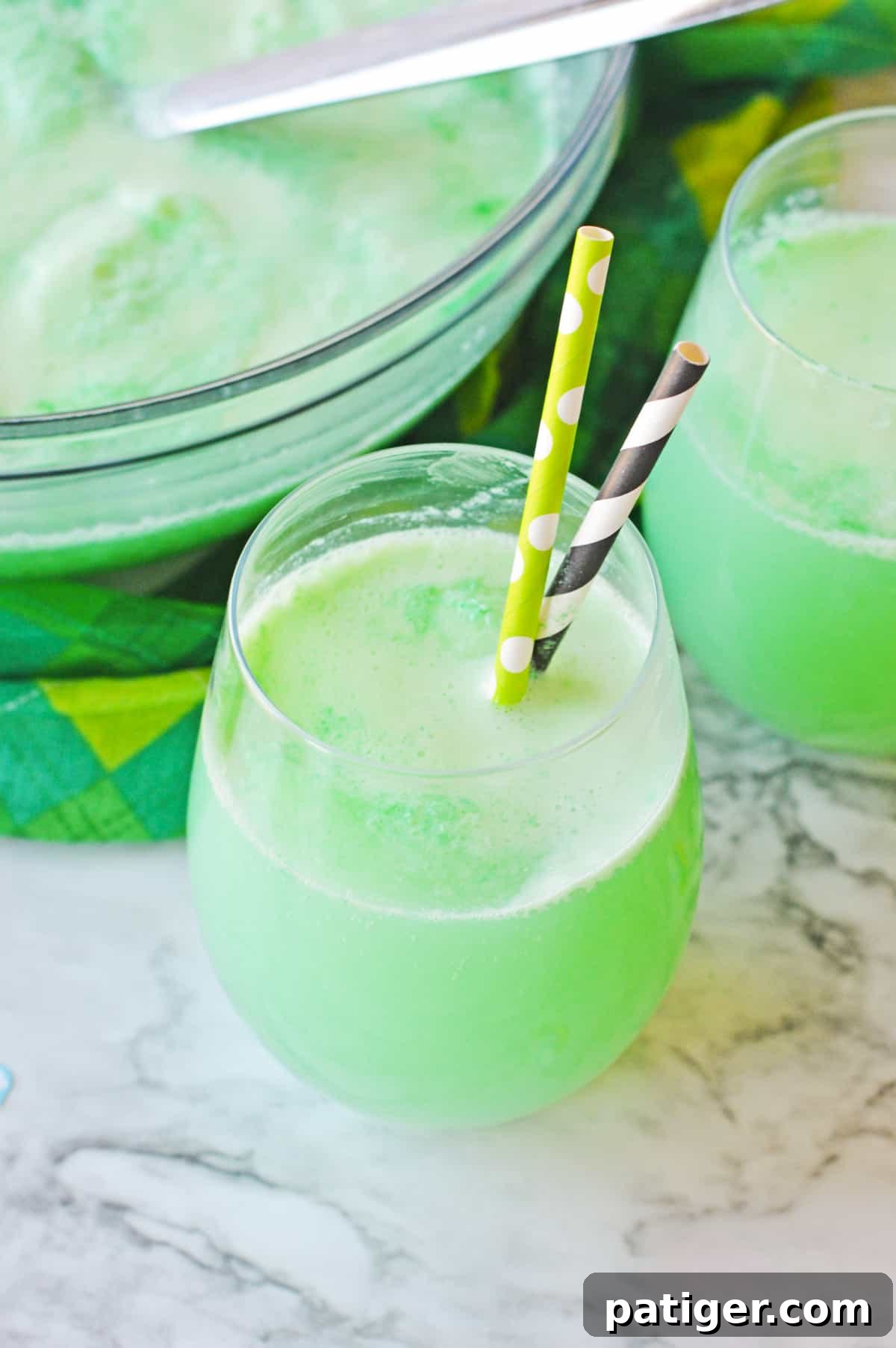 Clover Kick 6 St. Patrick's Day Alcoholic Punch in stemless wine glass with two paper straws and punch bowl filled with green punch behind it.