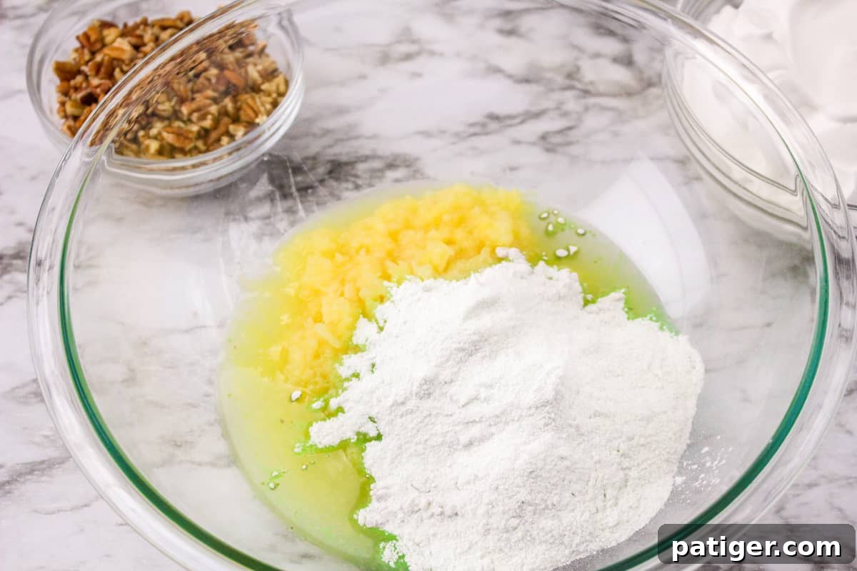 Crushed pineapple and pistachio pudding mix being stirred together in a clear glass mixing bowl, showing the initial blending of ingredients.