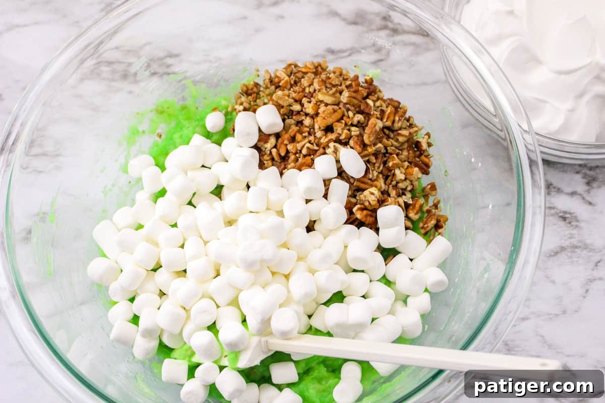 Mini marshmallows and chopped pecans added on top of the green pistachio pudding and pineapple mixture in a glass mixing bowl, ready to be stirred in.