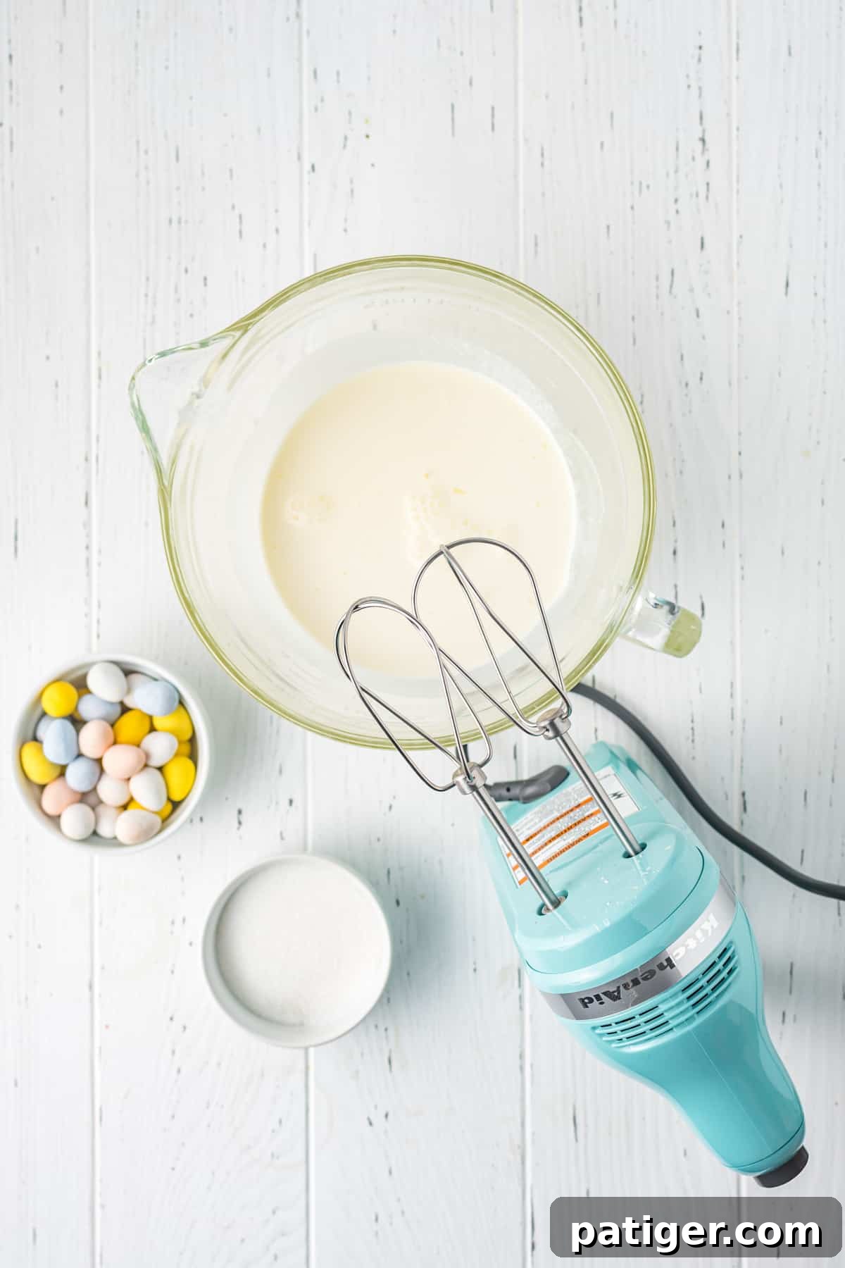 A hand mixer beating heavy whipping cream in a large glass bowl, creating a fluffy, light filling for the Easter cake roll.