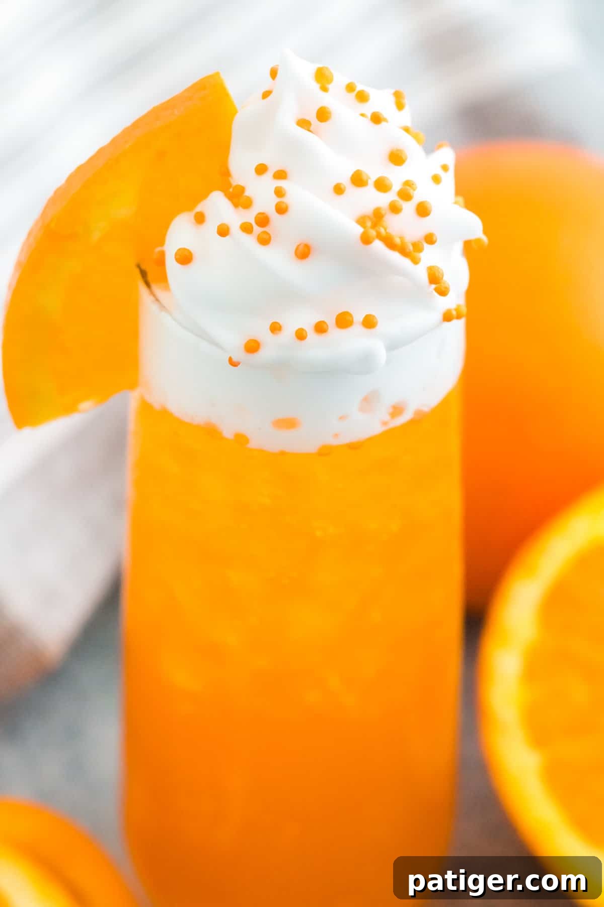 A tall glass of Orange Creamsicle Cocktail, topped generously with whipped cream, a fresh orange slice, and playful orange sprinkles. The drink itself is a vibrant orange hue, suggesting a refreshing and sweet experience.