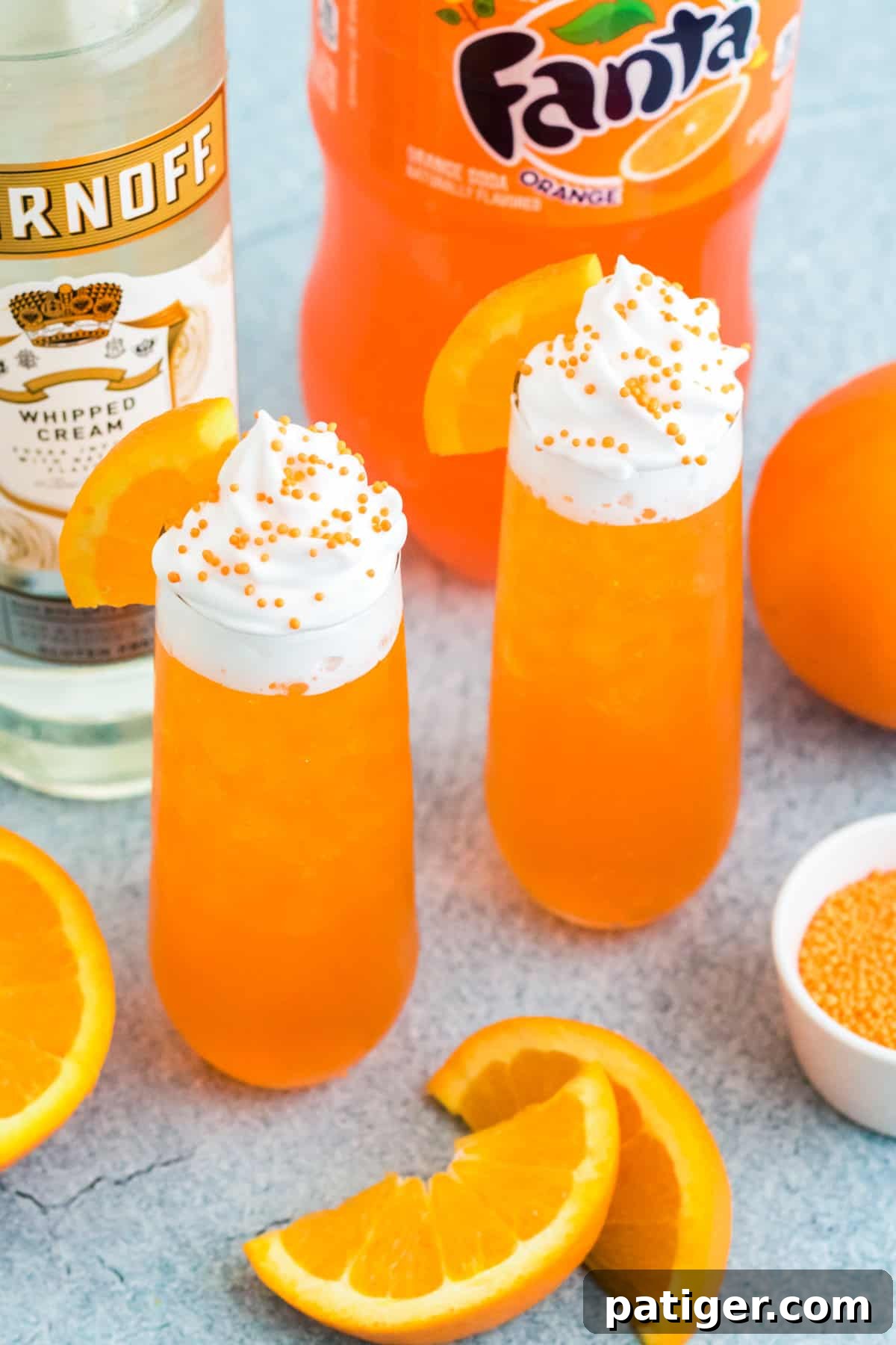 Two perfectly prepared creamsicle cocktail drinks, topped with whipped cream and garnished with fresh orange wedges. They are presented alongside a bottle of vodka and a can of orange soda, highlighting the simple ingredients used.