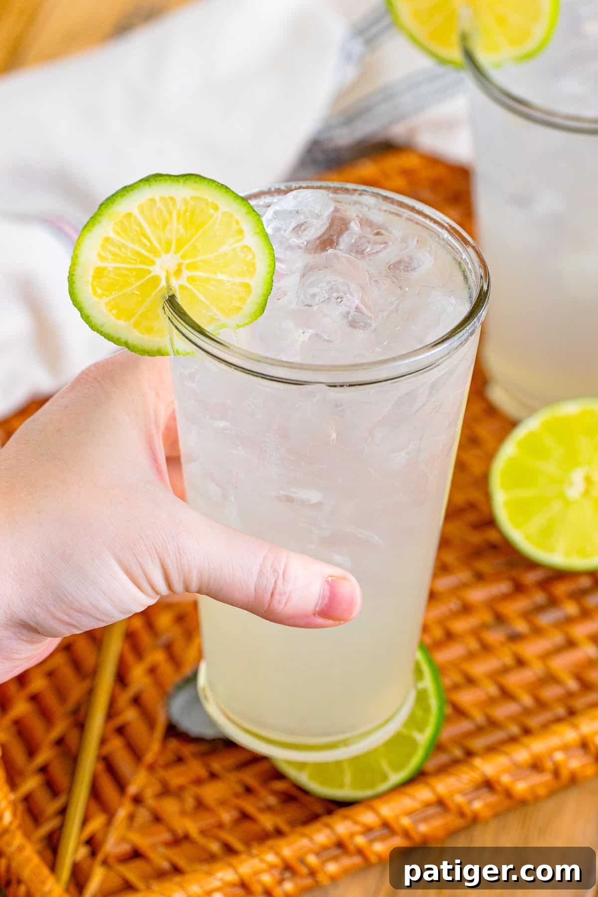 A hand holding a tall glass of Ranch Water cocktail, garnished with a lime wheel, against a bright background.
