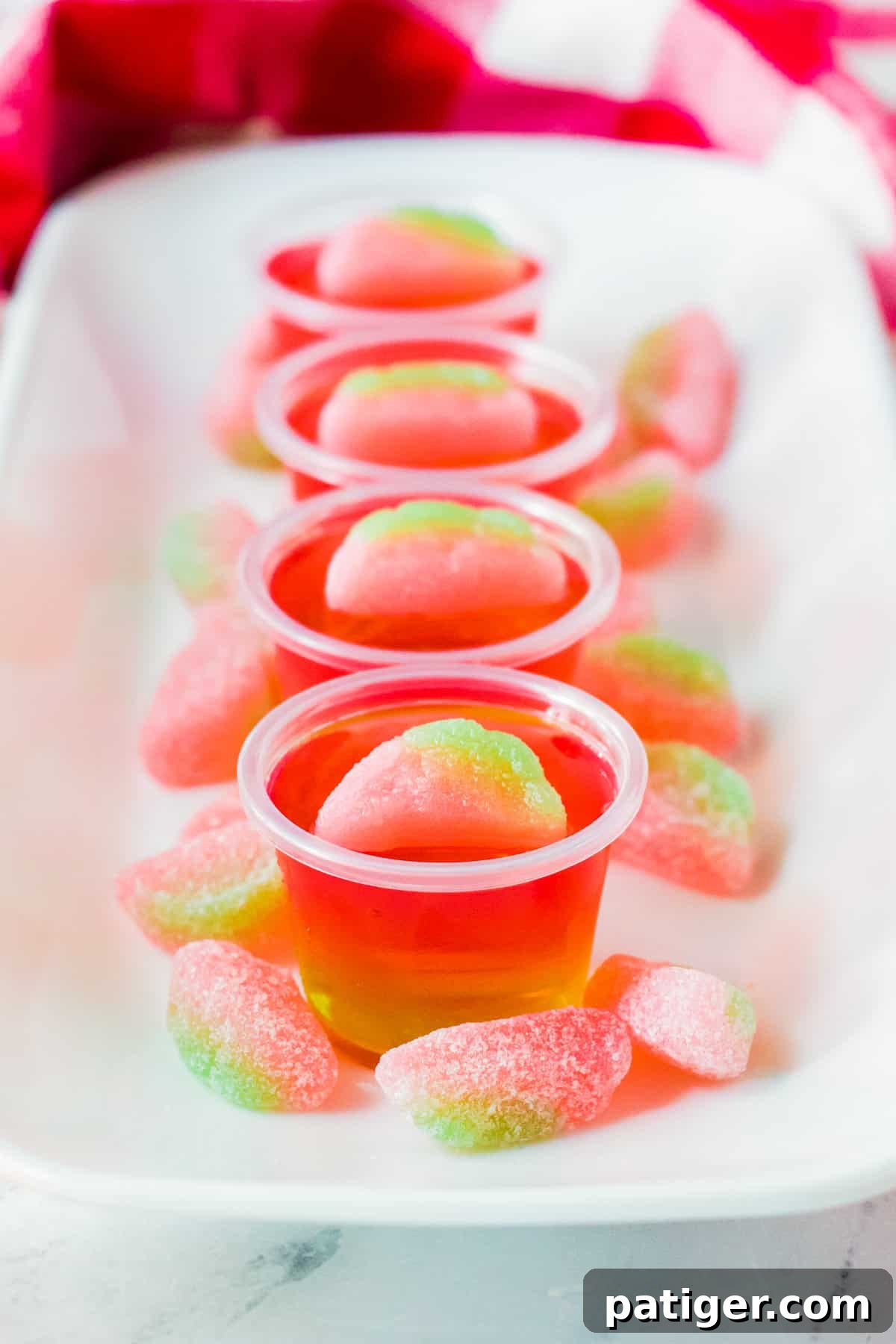 Vibrant pink and green layered watermelon vodka jello shots on a white serving tray, surrounded by extra sour watermelon candies.