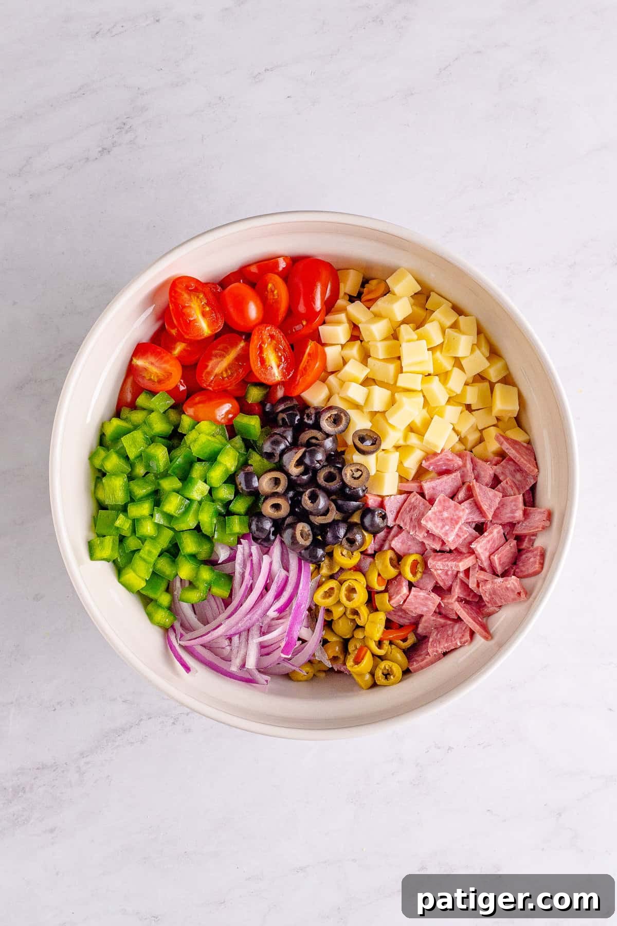 Large bowl filled with cooked rotini pasta, halved grape tomatoes, chopped green pepper, sliced red onion, sliced black and green olives, chopped salami, and cubed mozzarella cheese, ready for dressing.
