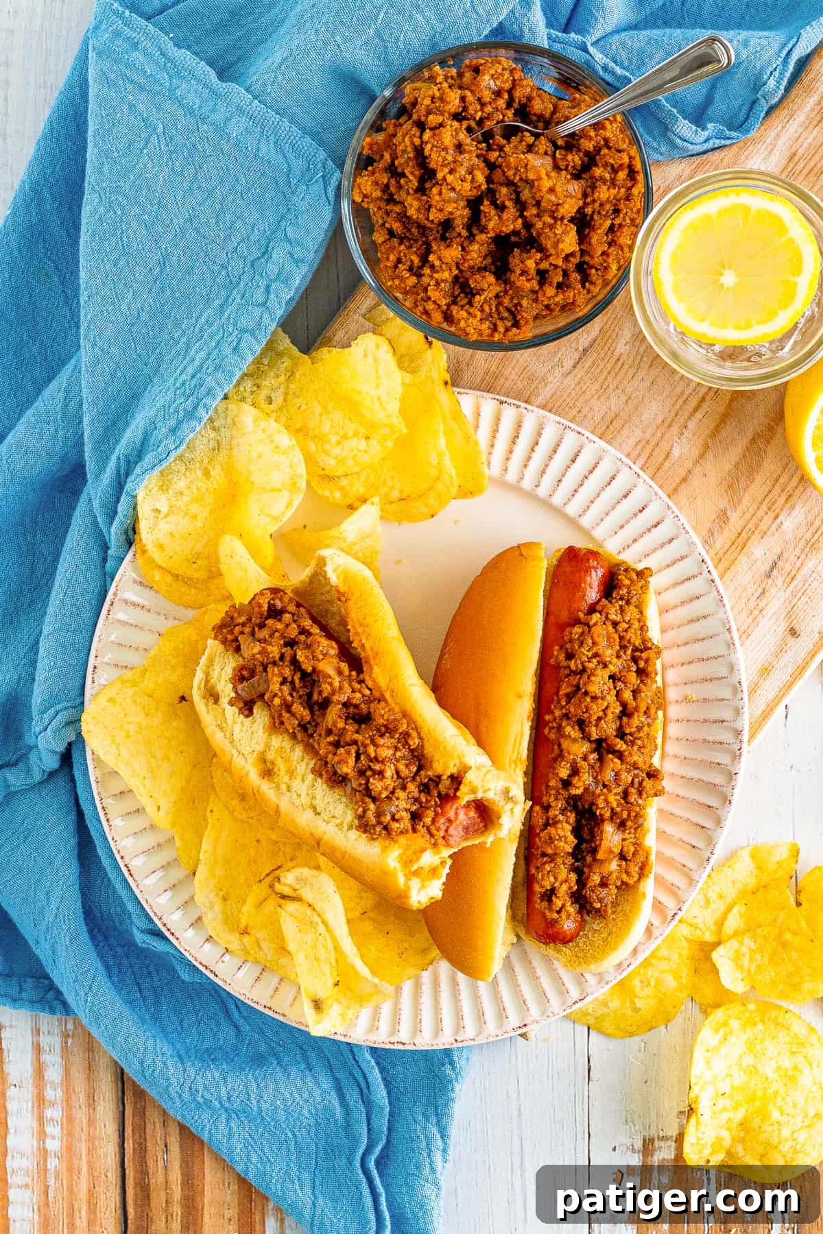 Hot dogs with chili sauce served with potato chips. More chili can be seen in background.