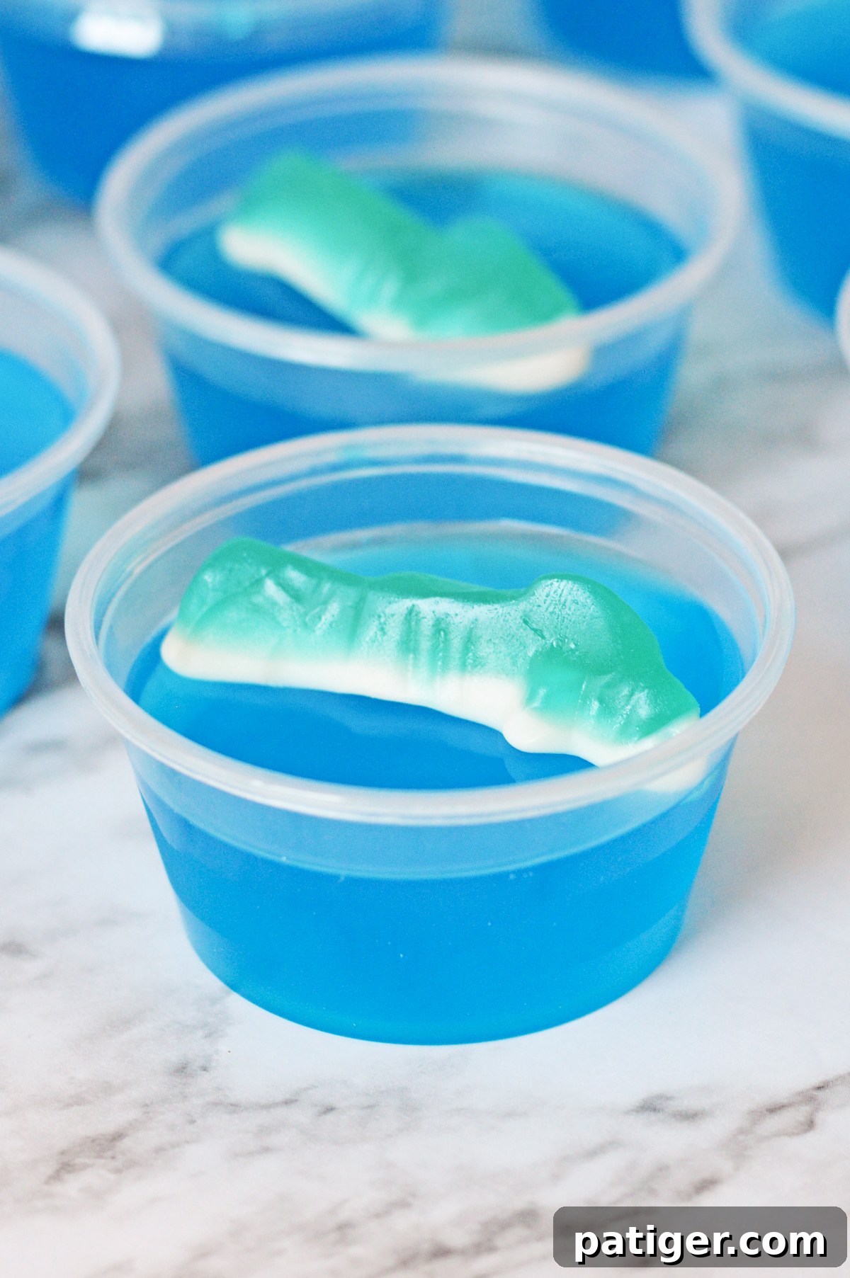 A single blue shark jello shot topped with a gummy shark candy, set against a blurred summer background.