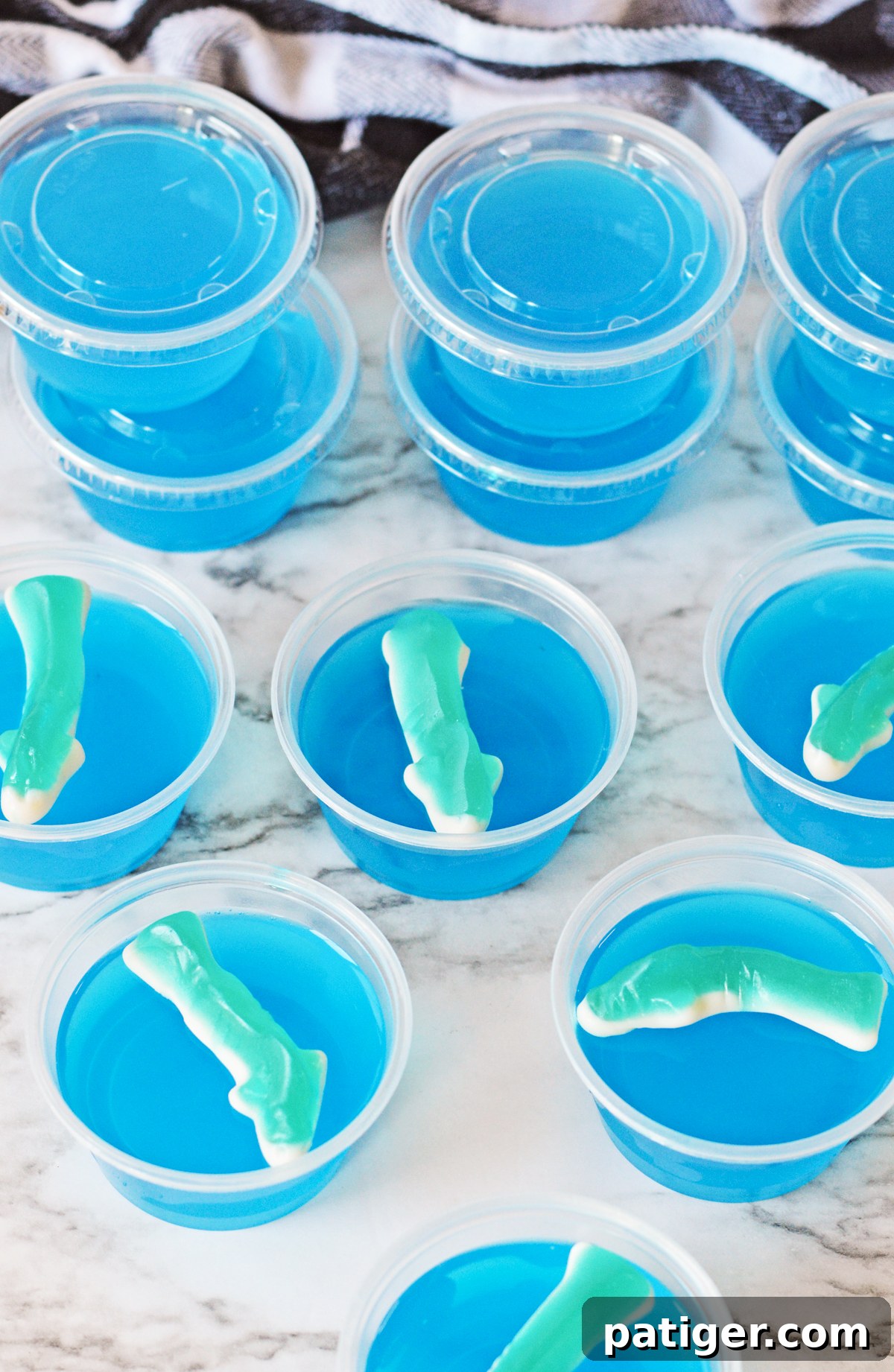 Multiple blue gummy shark jello shots in small plastic shot glasses, some with clear lids, arranged neatly.