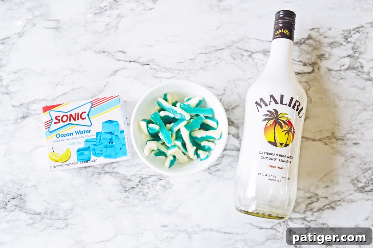 Ingredients for shark jello shots: a box of Sonic Ocean Water jello, a bowl of blue shark gummy candies, and a bottle of Malibu coconut rum.