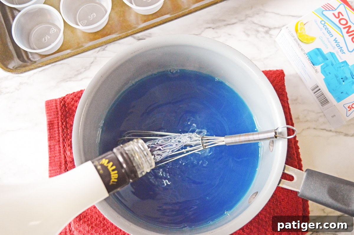 A bottle of Malibu coconut rum being poured into a blue jello mixture in a glass bowl to make shark shots.
