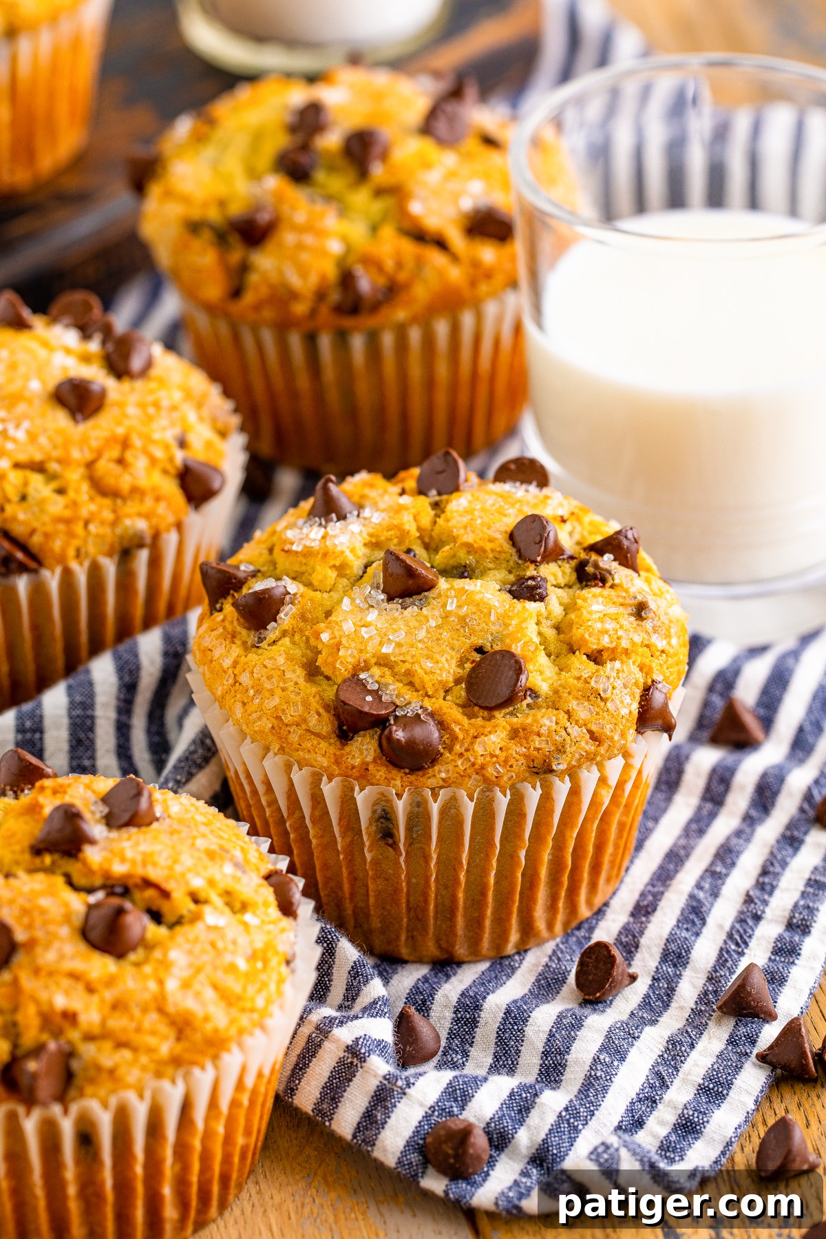 Ultimate Bakery Chocolate Chip Muffins 2 Freshly baked bakery-style chocolate chip muffins on a wooden table, accompanied by a glass of milk and a blue and white striped linen.