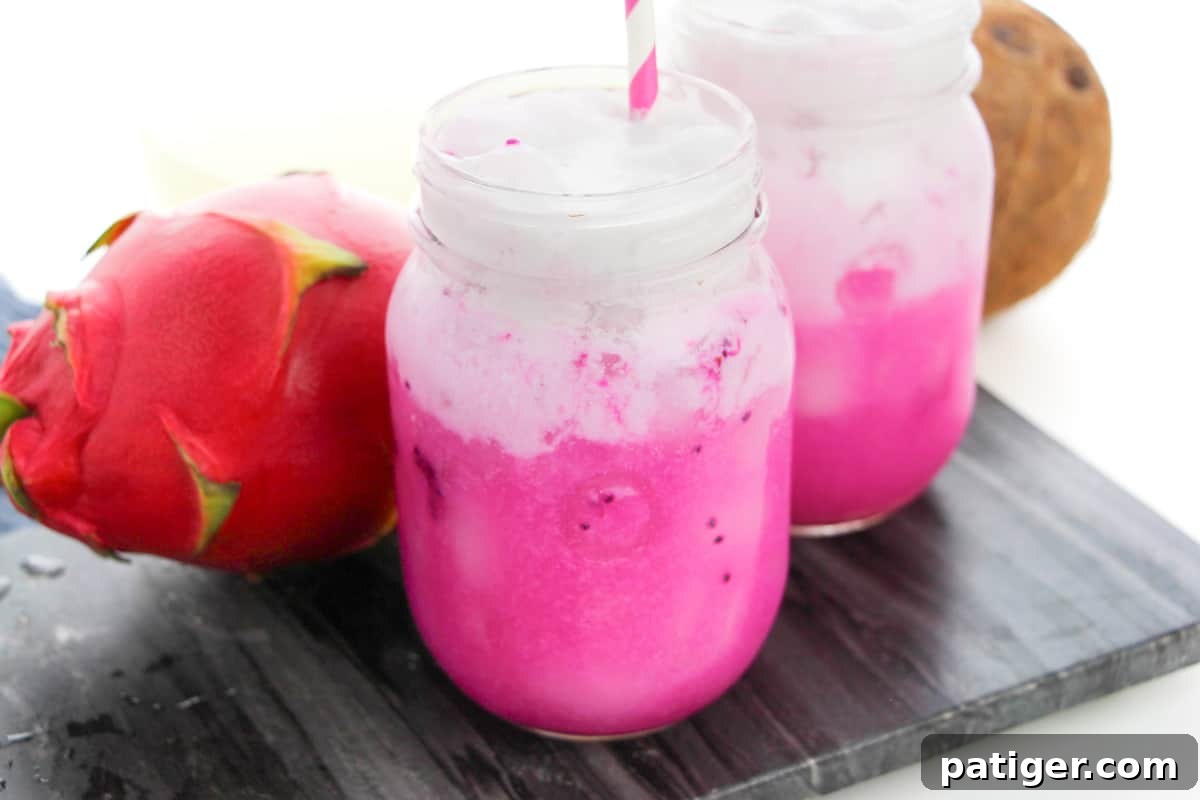 Copycat Starbucks Dragon Drink with vibrant pink mango dragonfruit refresher drink in mason jar topped with coconut milk. Another pink drink and a fresh mango and dragonfruit are in the background.