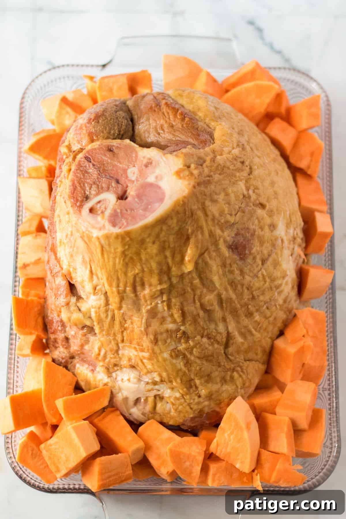 Bone-in spiral ham in a casserole dish surrounded by chopped sweet potatoes, ready for glazing.
