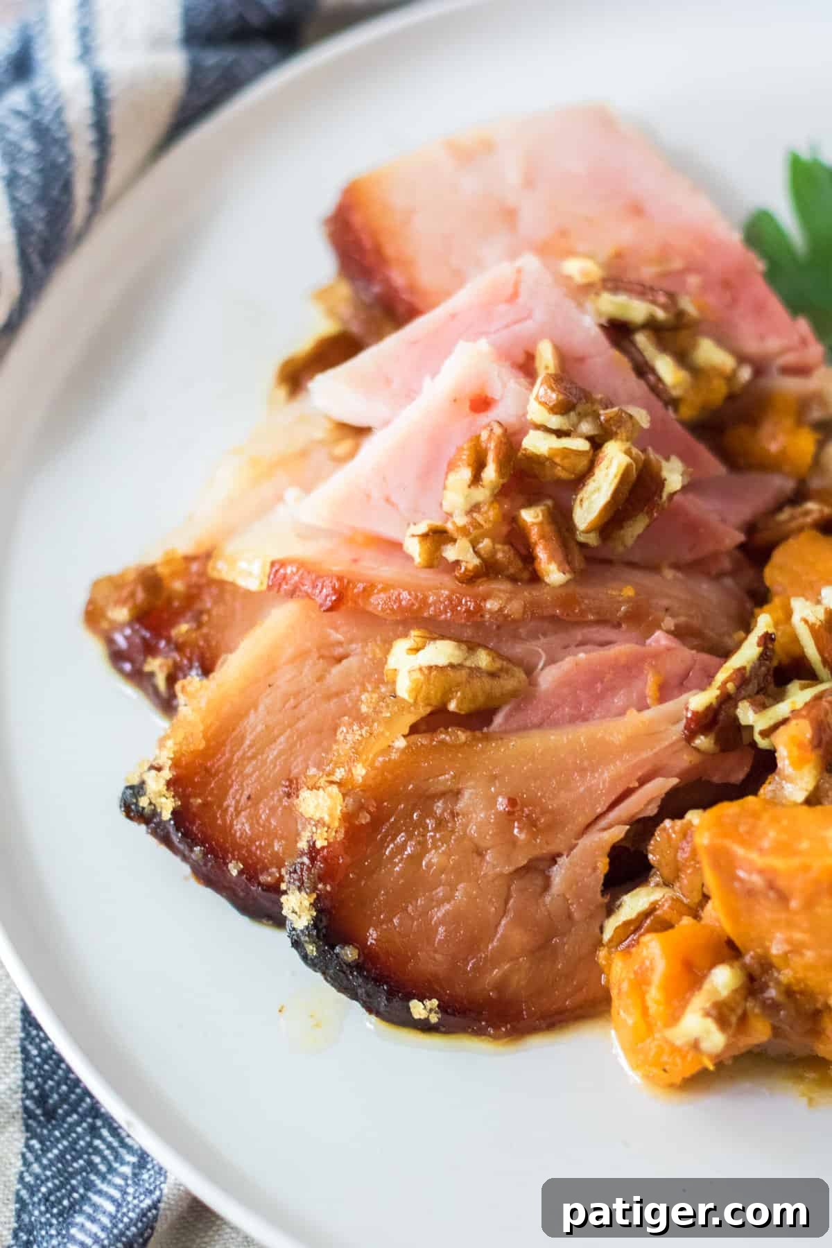 Slices of glazed spiral ham topped with pecans on a white plate, accompanied by sweet potatoes.