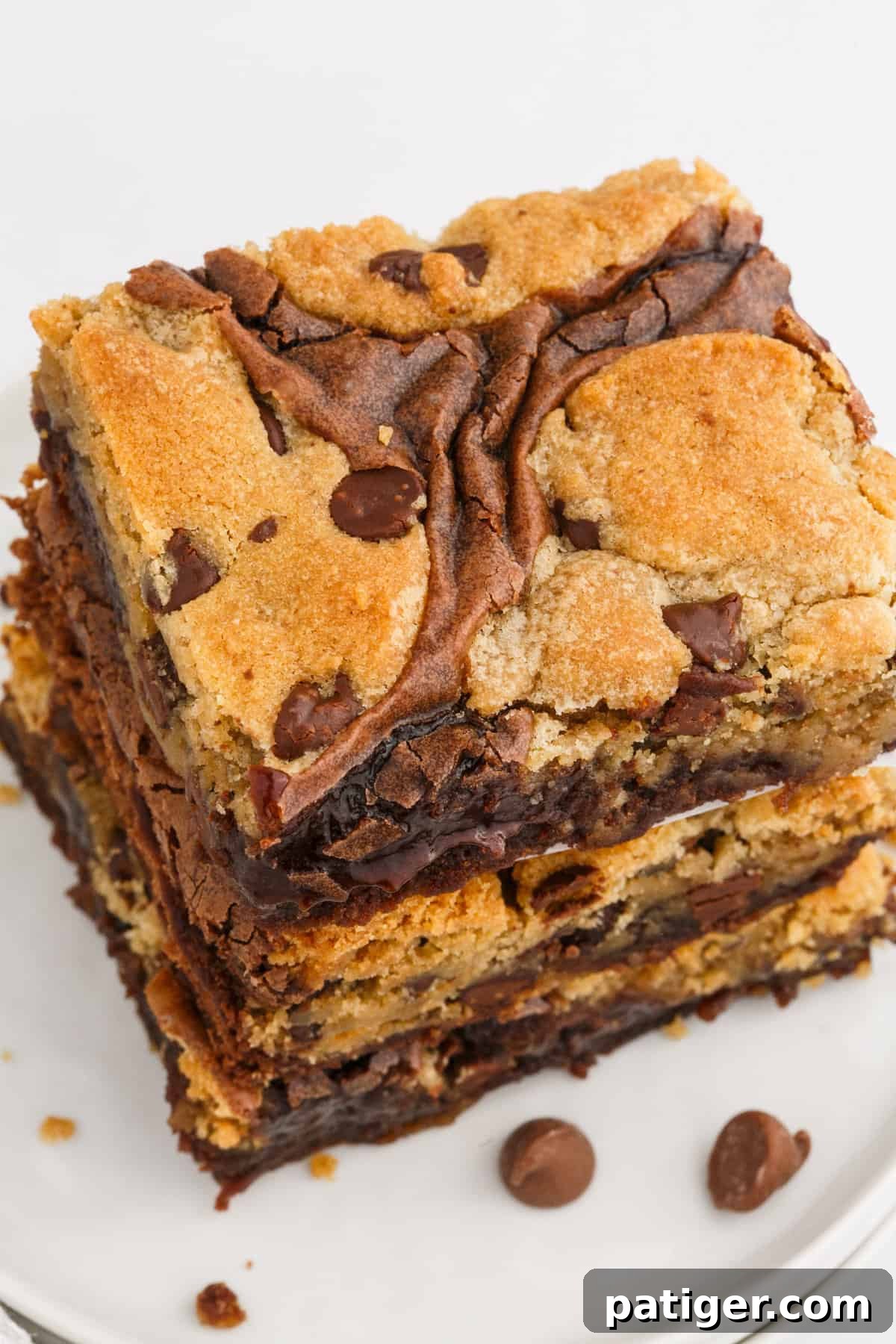 A close-up shot of a single chocolate chip cookie brownie bar on a white plate, with loose chocolate chips scattered nearby.