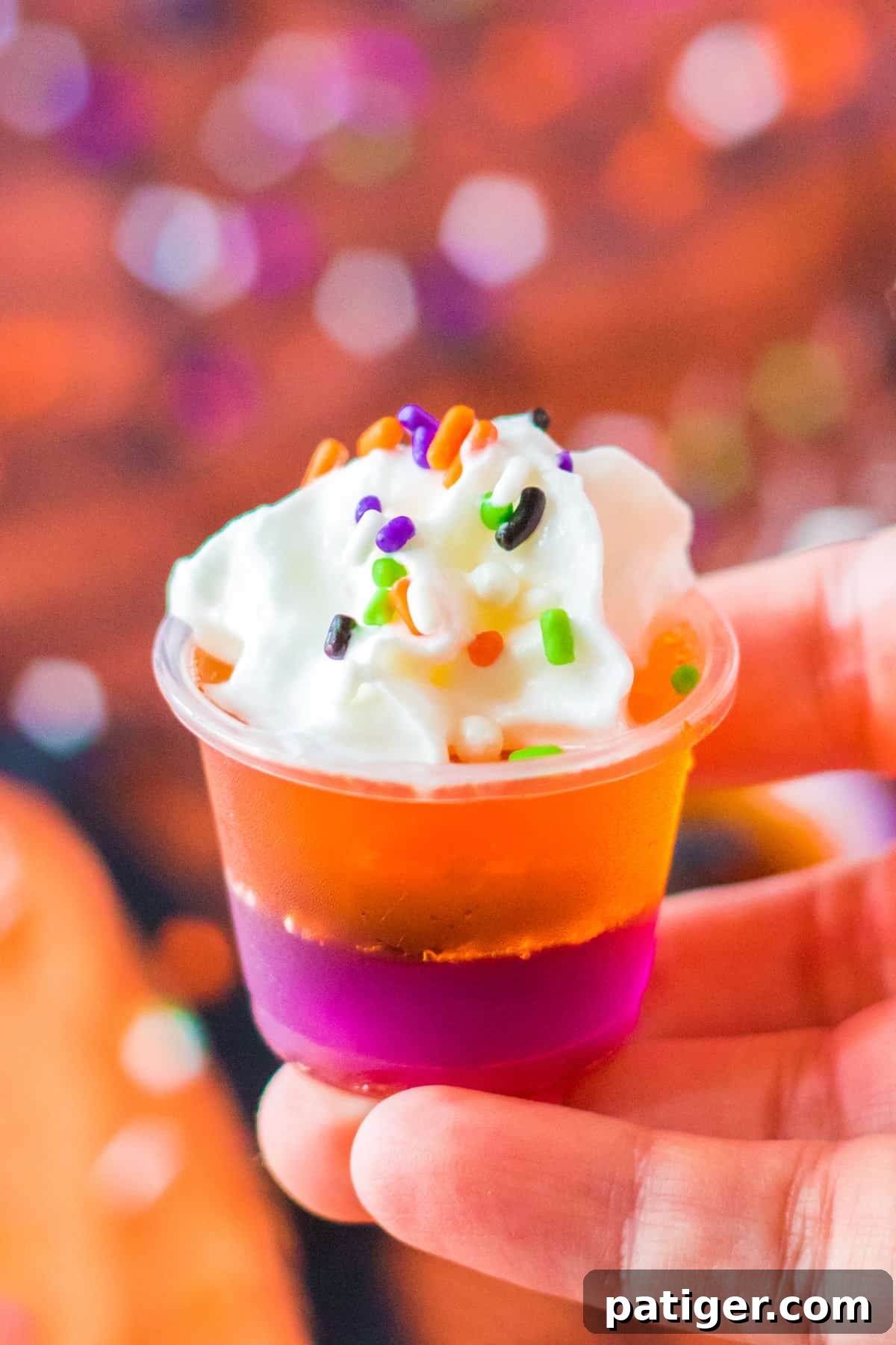 A hand holding a perfectly layered Halloween jello shot, garnished with whipped cream and sprinkles, showcasing its festive appeal.