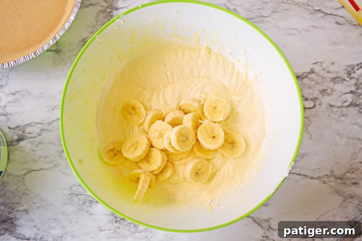 Freshly sliced bananas being gently folded into the banana pudding pie mixture in a mixing bowl.