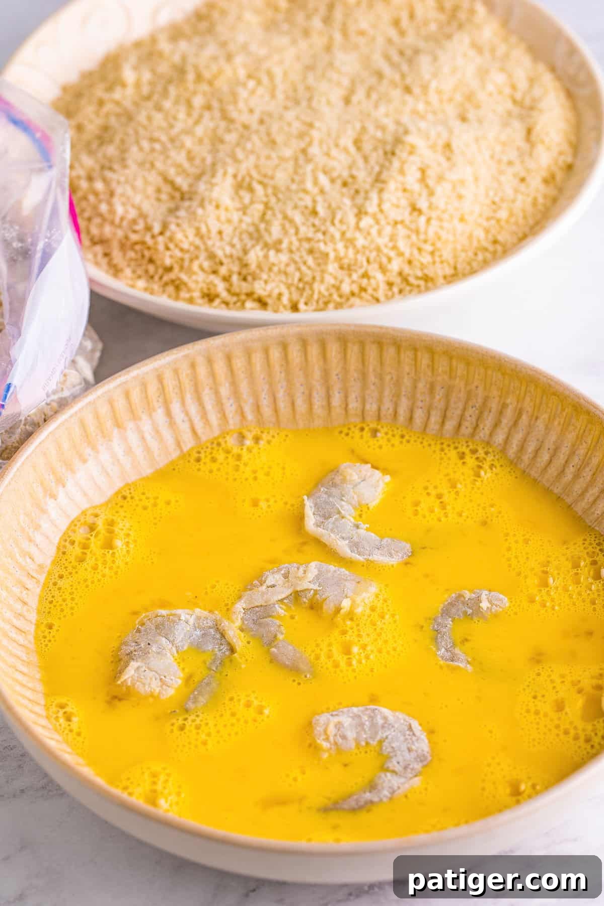 Several shrimp being dipped and coated in the egg wash mixture in a shallow bowl.