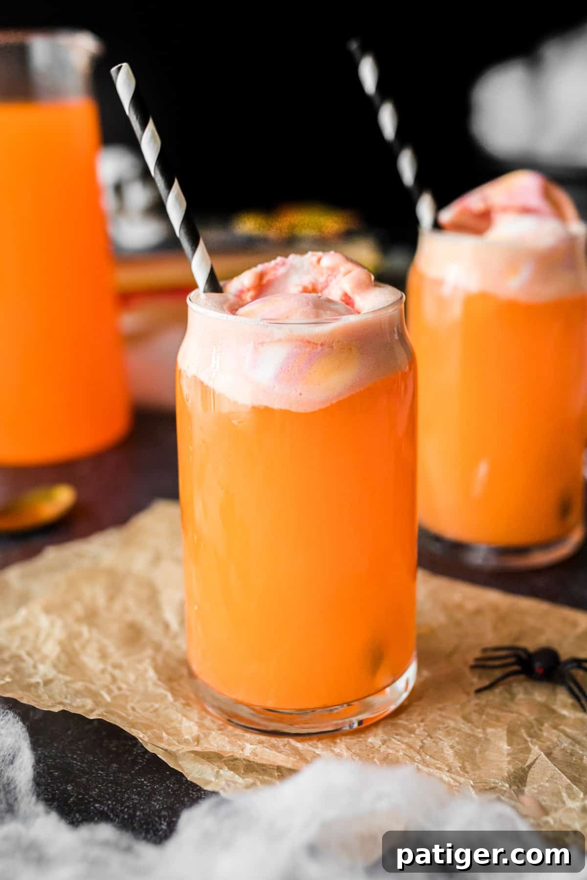 Whimsical Concoction 2 Orange Halloween punch in tall glass topped with sherbet. Another filled glass and pitcher of the punch are in the background.