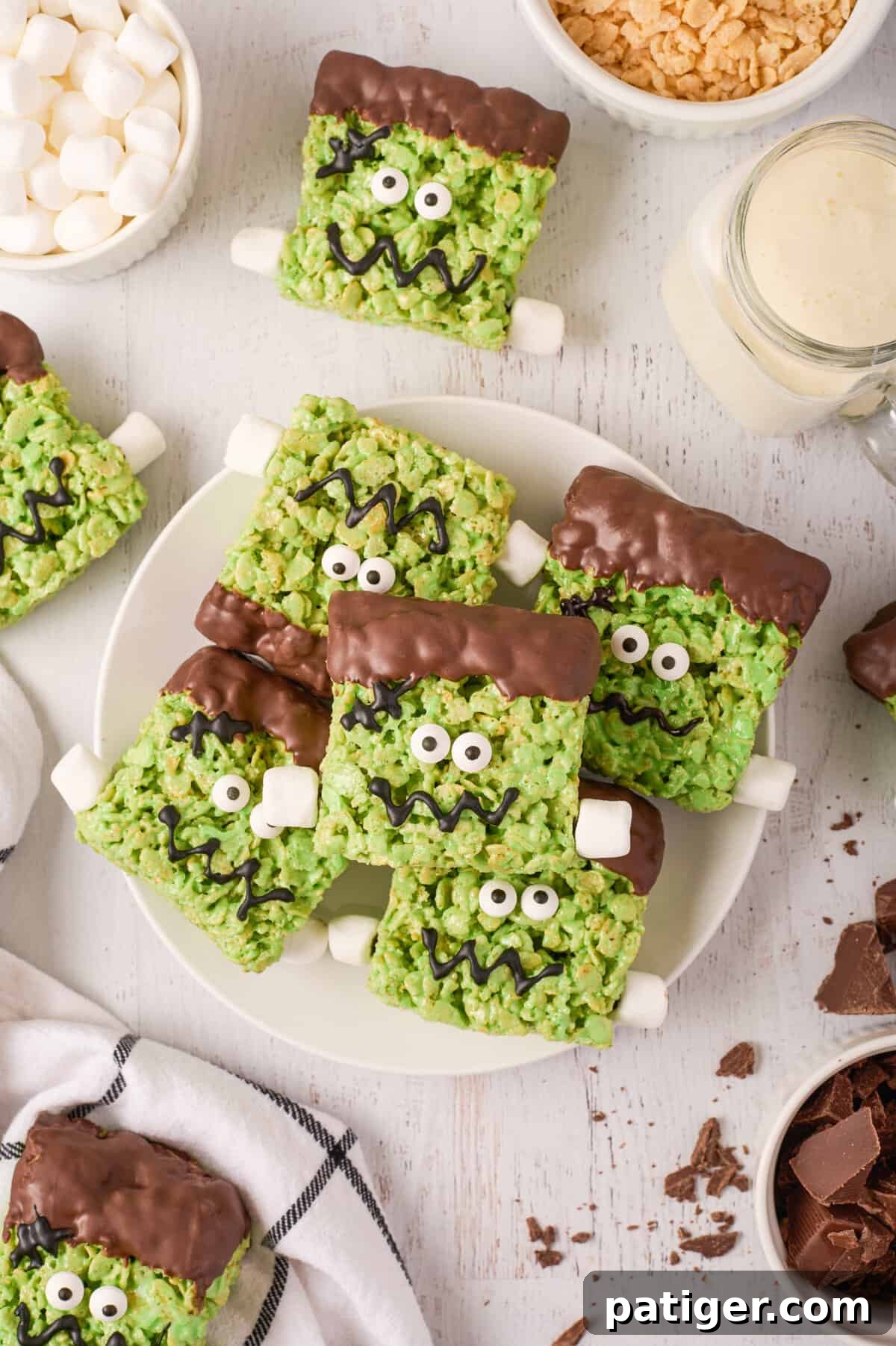 Spooktacular Frankenstein Krispies 14 Frankenstein treats piled on white plate with mini marshmallows, chopped chocolate, and other ingredients around them.