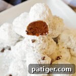 Double chocolate snowball cookies stacked on a white plate with one cookie cut in half to show chocolate interior packed with mini chocolate chips.