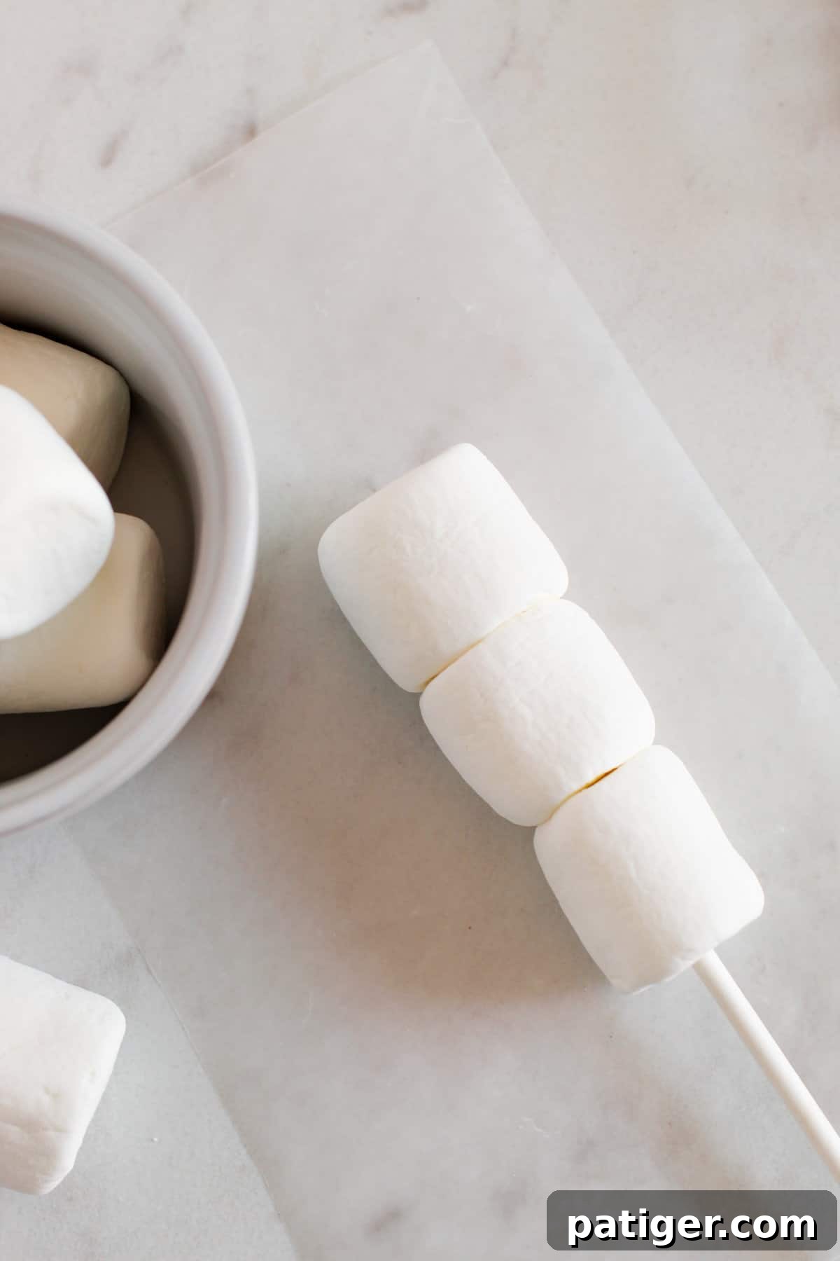 Three white marshmallows threaded onto a white treat stick, forming the base of a snowman.