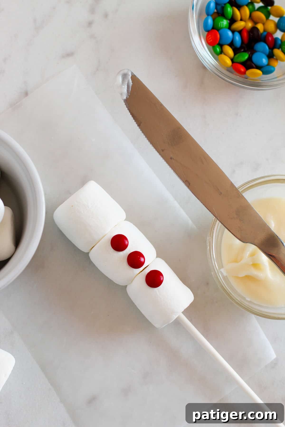 Three vibrant red M&M candies placed as buttons on a snowman pop, with a butter knife and icing on the side.