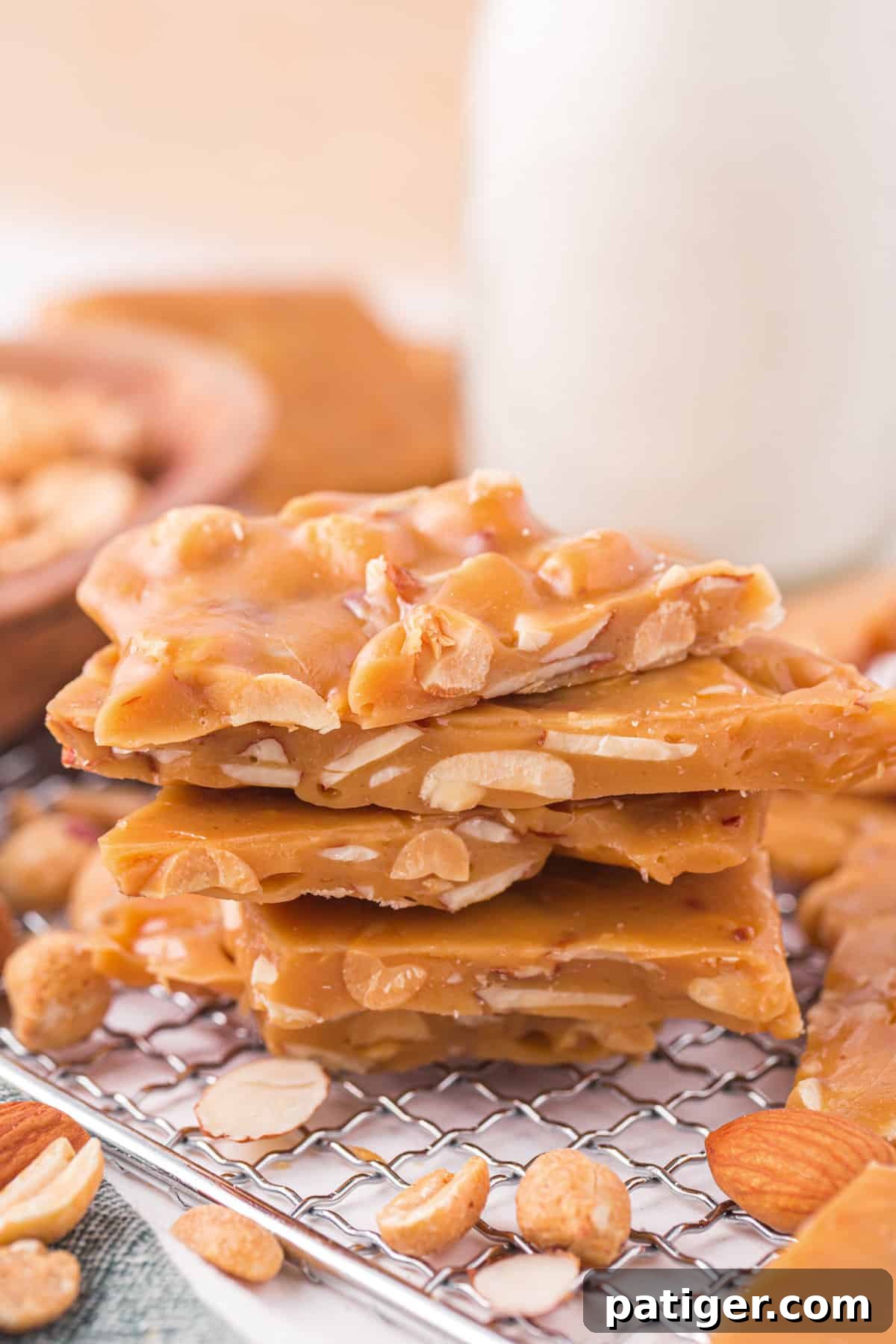 A close-up shot of golden, homemade nut brittle with visible chunks of almonds and peanuts, stacked artfully on a metal cooling rack.