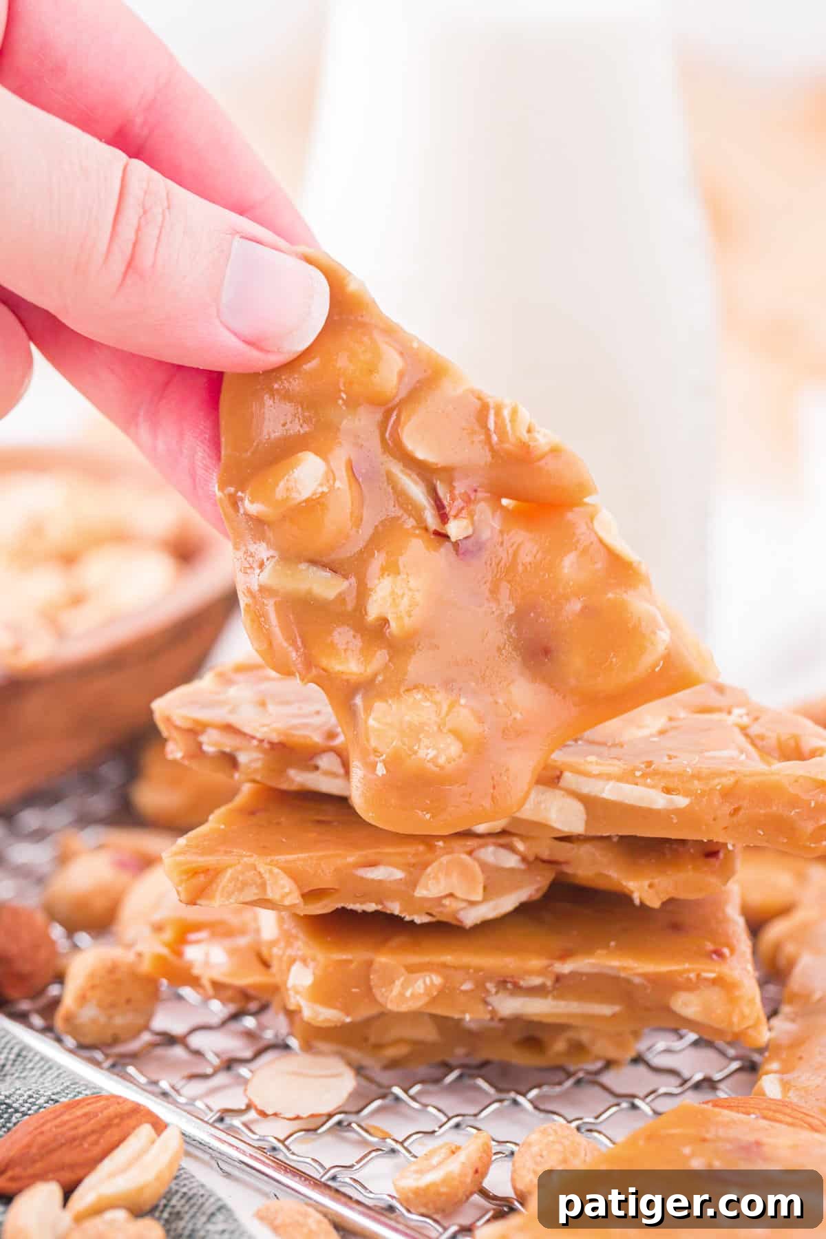 A hand gently holds a piece of golden caramel nut brittle, showcasing its jagged edges and the embedded nuts.