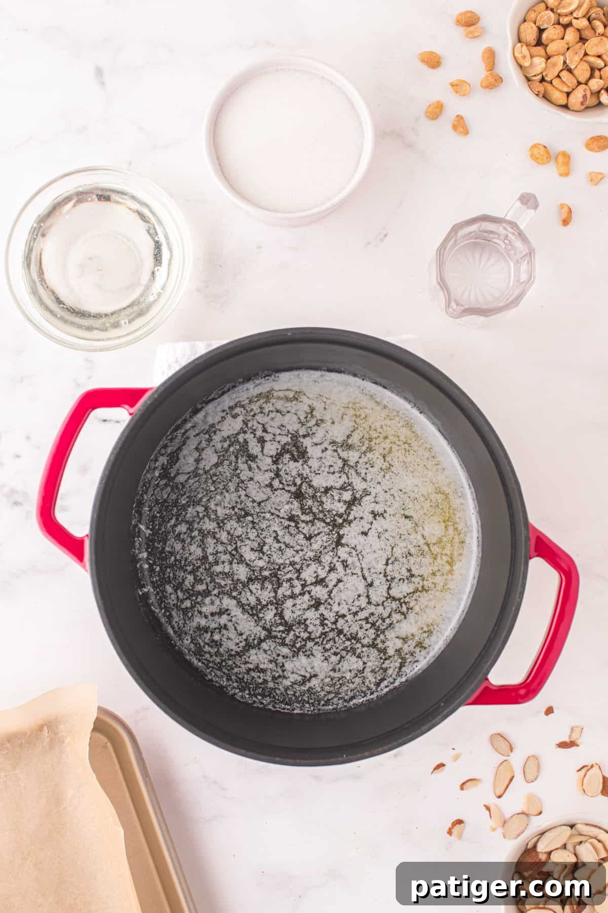 A large, heavy-bottomed pot on a stovetop with melted butter. Bowls of sugar, light corn syrup, water, baking soda, and mixed nuts are arranged next to it, ready for use.