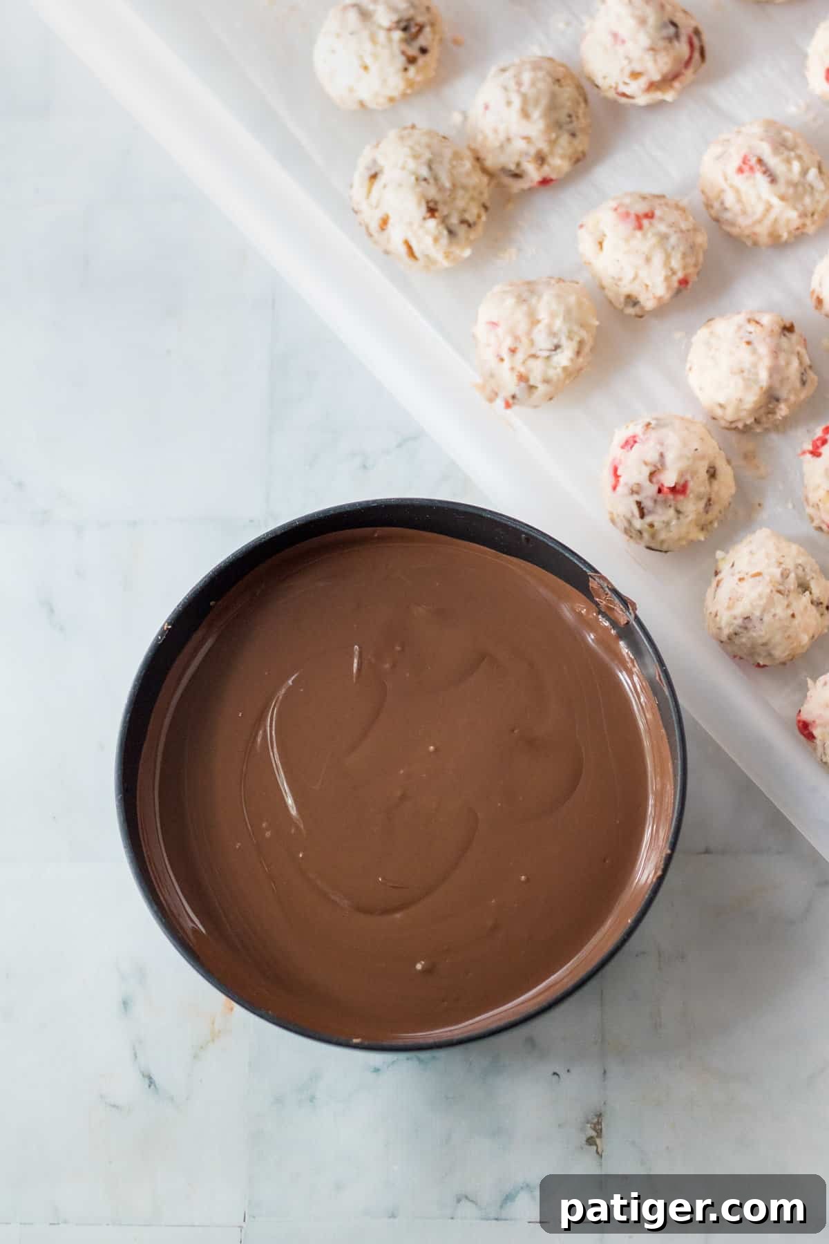 A small bowl of smoothly melted chocolate almond bark positioned next to a baking sheet filled with chilled, undipped Martha Washington candies.