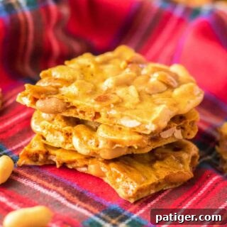 Pieces of peanut brittle stacked on top of one another with roasted peanuts sprinkled around them.