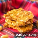 Pieces of peanut brittle stacked on top of one another with roasted peanuts sprinkled around them.